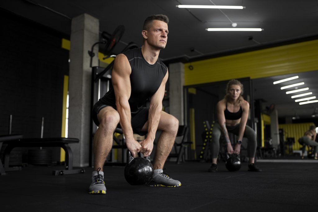 Two individuals performing kettlebell exercises in a gym, highlighting the importance of a gym buddy for motivation and promoting shared fitness activities.