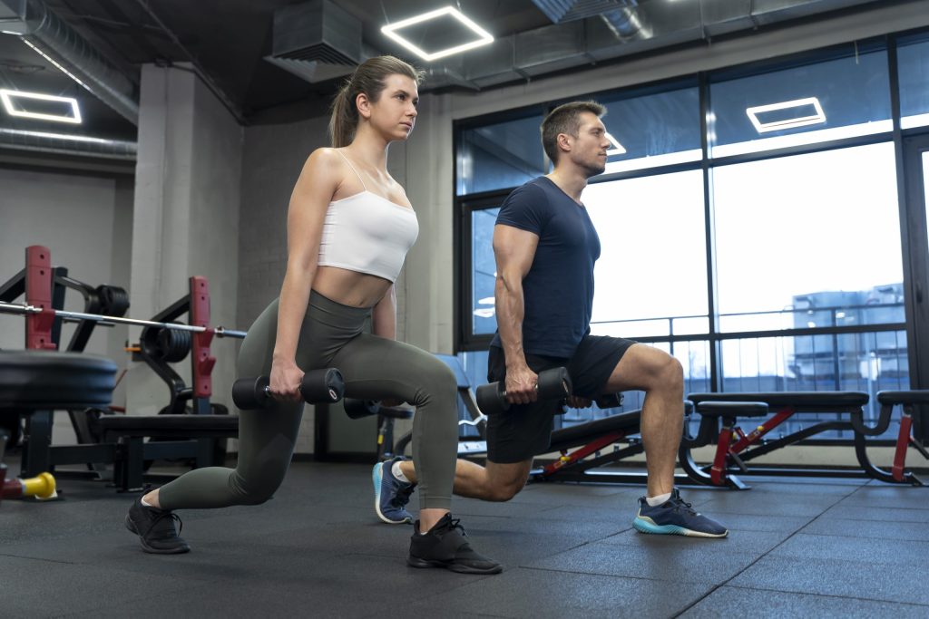 A man and a woman performing dumbbell lunges in a gym, emphasizing the importance of having a gym buddy to stay motivated and engage in effective fitness activities.