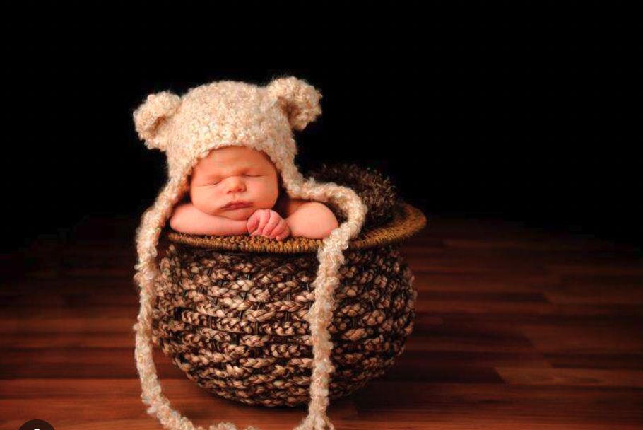 A newborn baby peacefully sleeping in a knitted basket, wearing a bear-themed hat, representing the creative collaboration and skill-building opportunities that come with having a photography buddy.