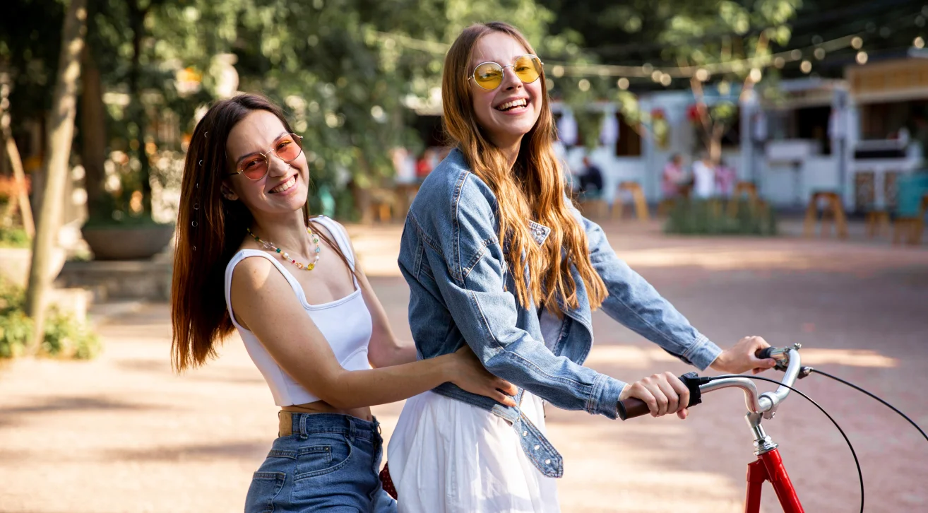 Two women enjoying a sunny day outdoors, smiling and posing with a bicycle in a lively community setting. A perfect depiction of making friends as an adult while engaging in fun activities. Platforms like Bunchups make it easy to connect through shared hobbies and interests in small gatherings.