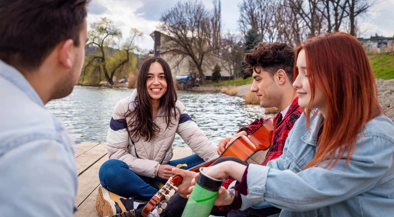 A group of friends sitting by a serene lakeside, playing guitar and sharing smiles, symbolizing friendship and healing after loss.